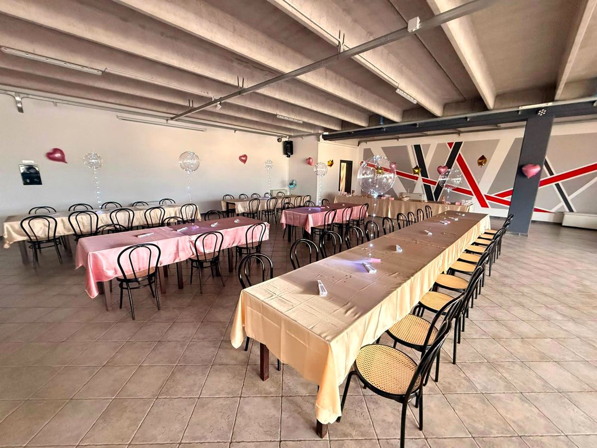 Salone allestito a banchetto: tavolate lunghe con tovaglie e sedie bistrot in stile Thonet, sullo sfondo la parete a righe rosse e grigie