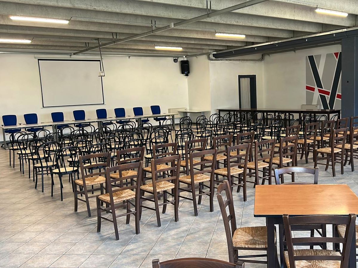 Salone in configurazione a platea: file di sedie, tavolo relatori e schermo per le proiezioni